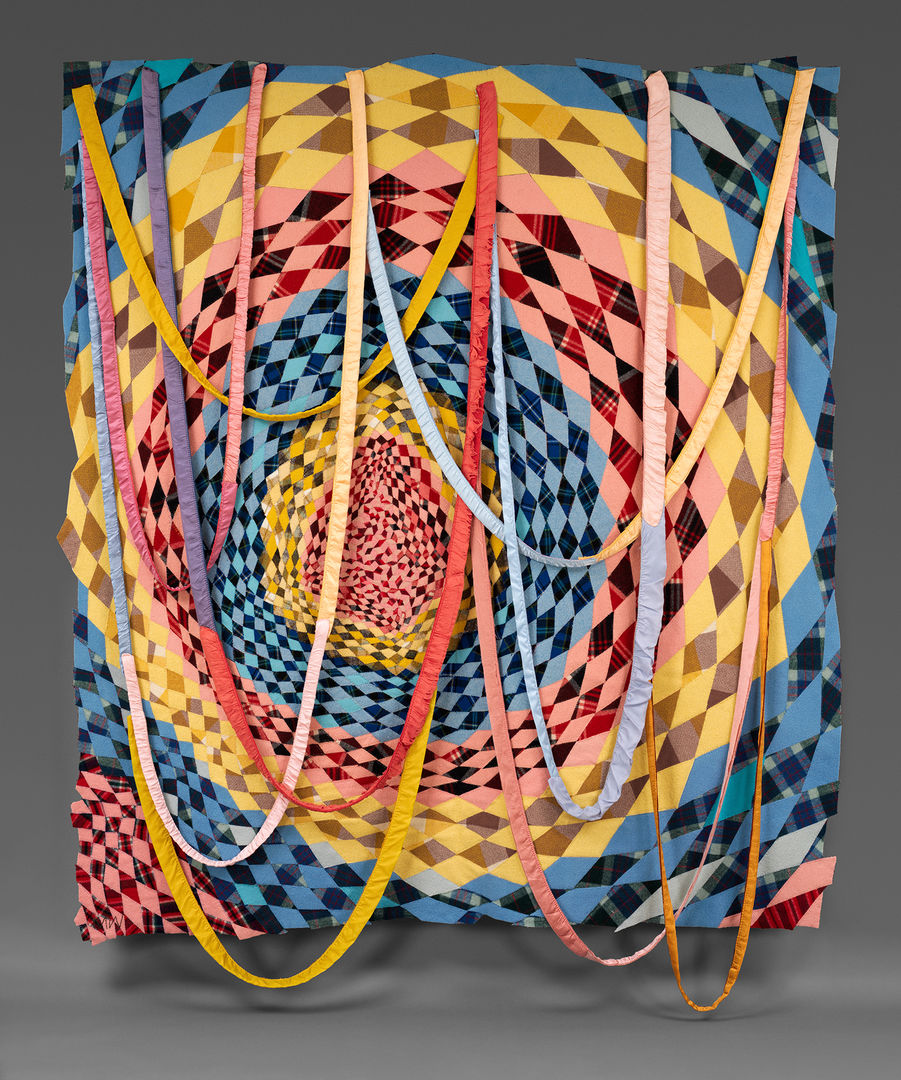 An abstract dreamcatcher. Blue, yellow, and red diamond shapes are quilted in a circular pattern, while long, thin strips of fabric are draped in loops over the frame.