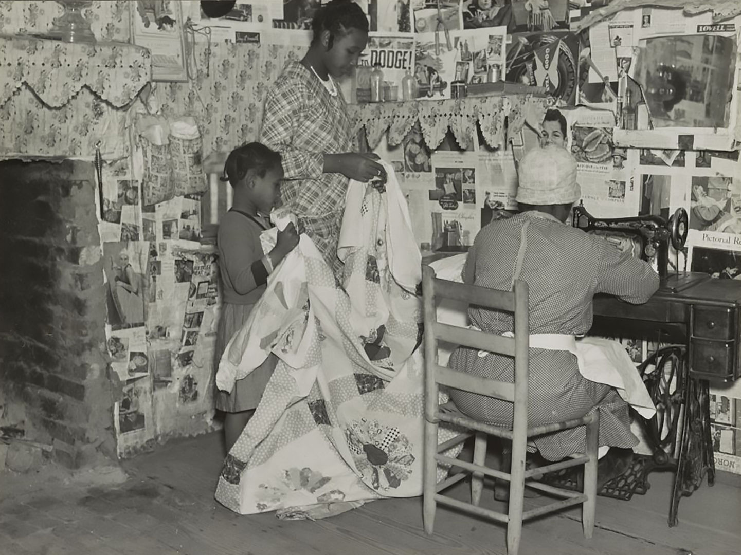 Black-and-white photograph of three people in a room decorated with wallpaper and advertisements. Two children hold up a white quilt being worked on by a figure at a sewing machine.