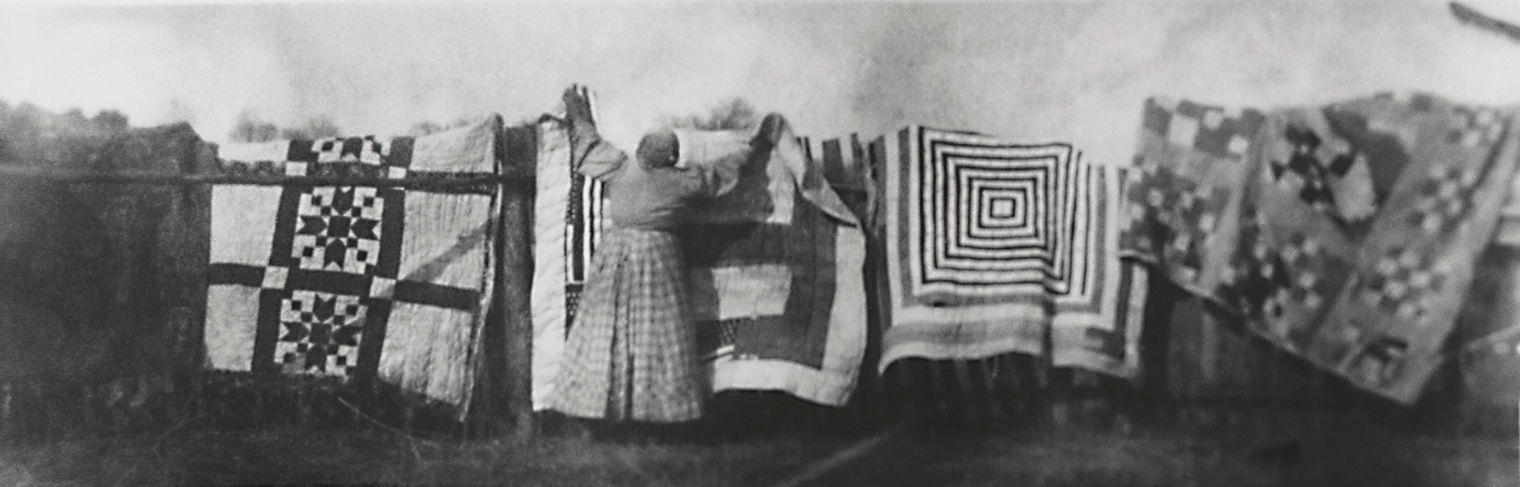 Fuzzy black-and-white photograph of a woman hanging quilts outdoors.