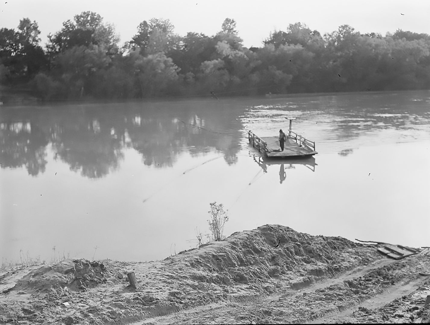 Black-and-white photograph of a river crossing. Trees cover the far side of the bank, while dirt marks the near side. In the water, a single figure crosses on a cable ferry.