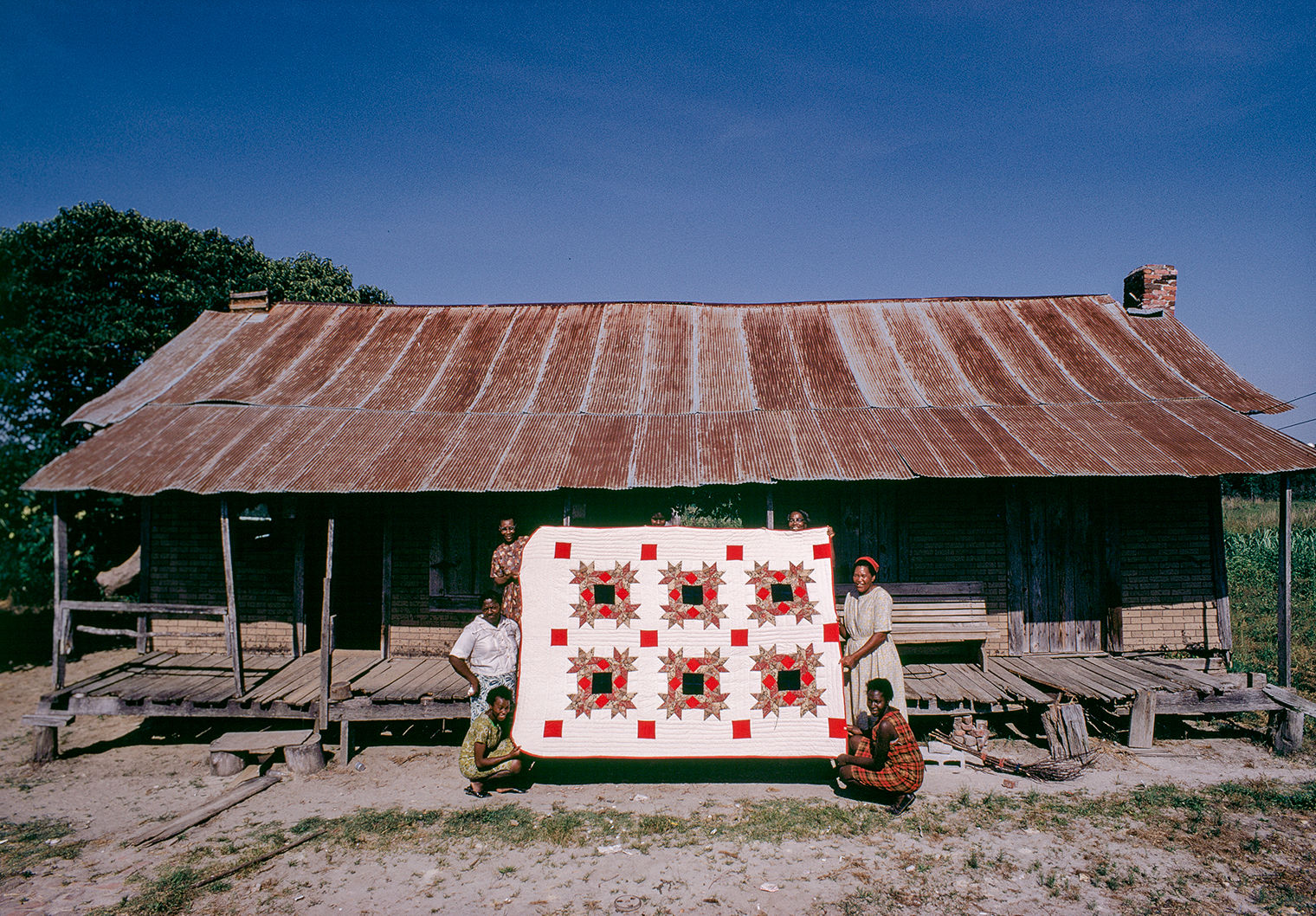 Photograph of seven women holding a red, white, and black patterned quilt. They are posing outside in front of a brick house with a sloping roof of corrugated metal.