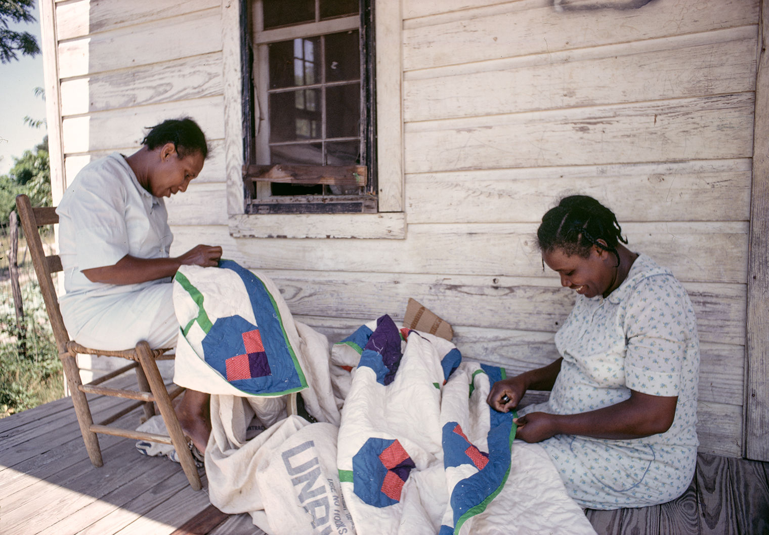 Two women smile while quilting outside on a porch. One is seated in a chair, while the other sits on the ground.