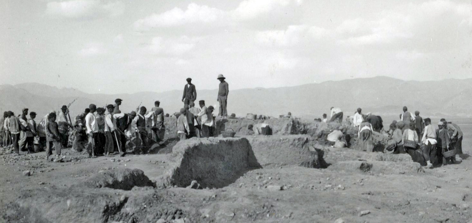 Black-and-white archival image of dozens of people excavating at Nishapur. The environment is primarily dirt, with mountains in the distance.