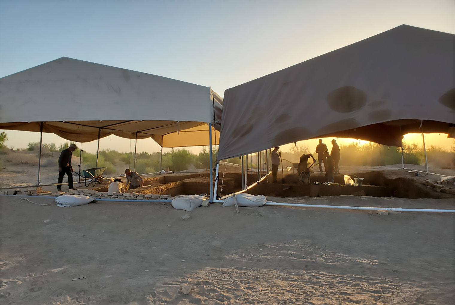 Image of excavation in progress at Dandanakan. Two large tents cover workers digging. The environment is scrubby, and the sun is low.