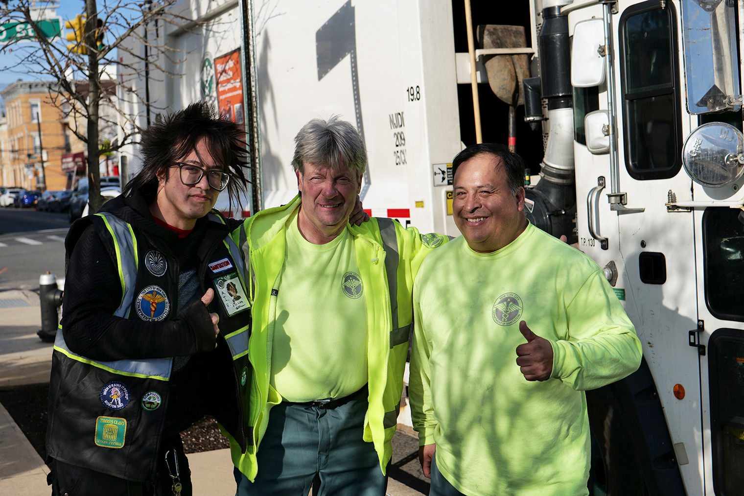 Photograph of three people in black and bright green Sanitation Department attire. They are posing outside in front of a box truck.