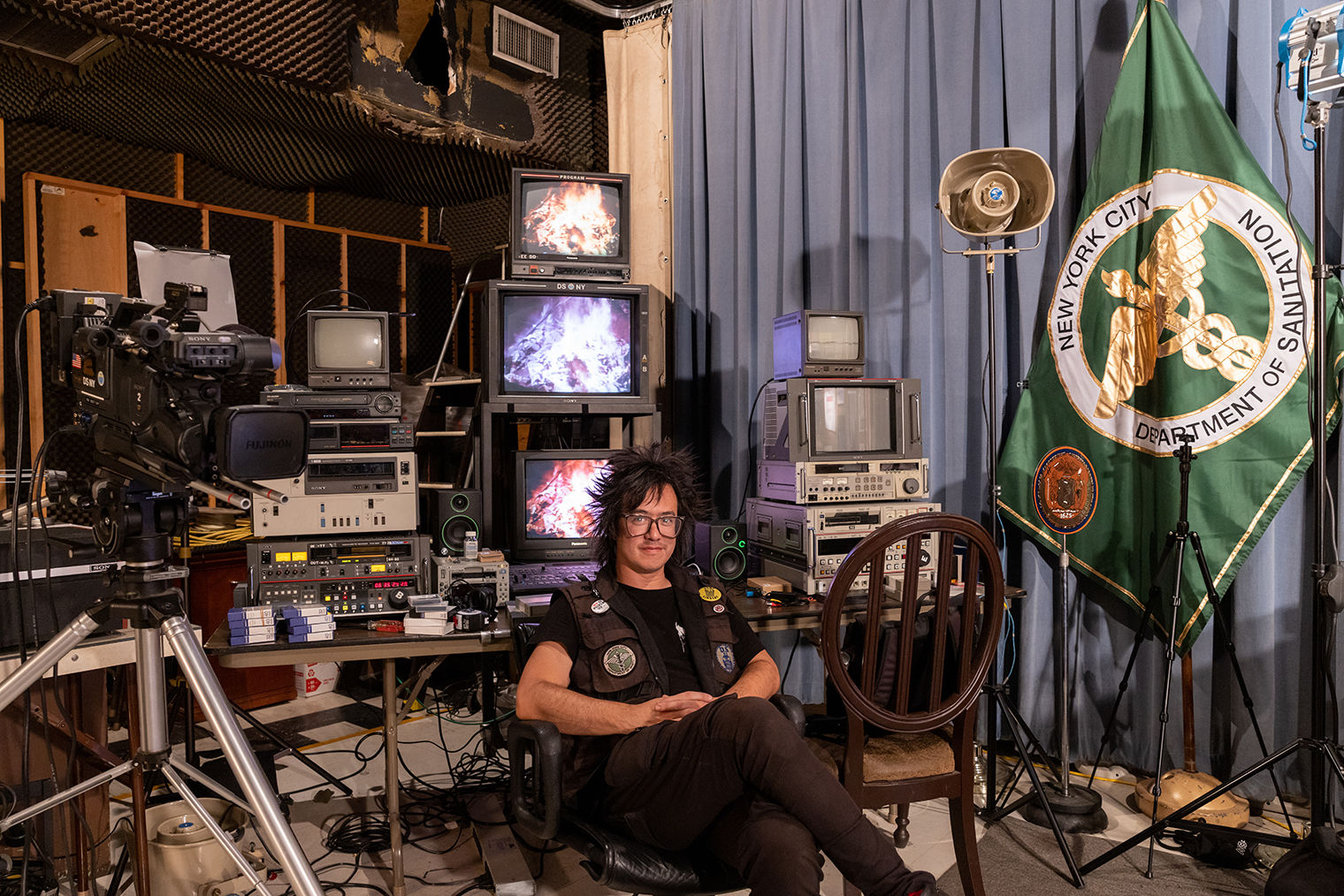 The artist sTo Len posing in front of an array of old TVs and other dated electronic equipment in a studio-like space.