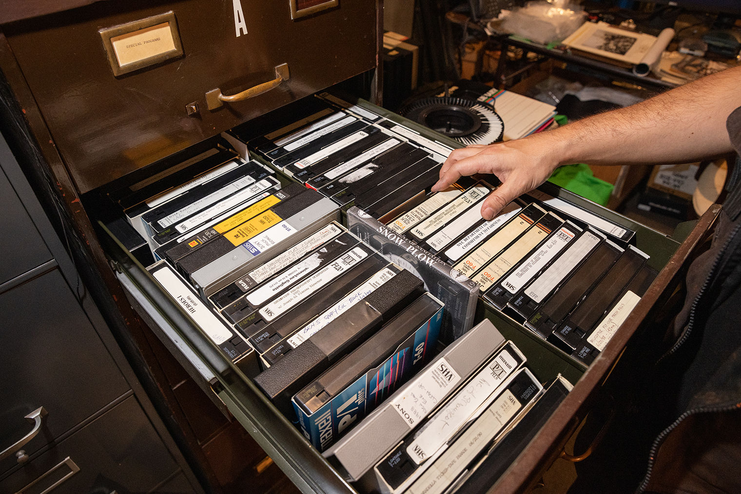 A file cabinet drawer opened to reveal many VHS tapes. A hand is reaching at a group of three tapes.