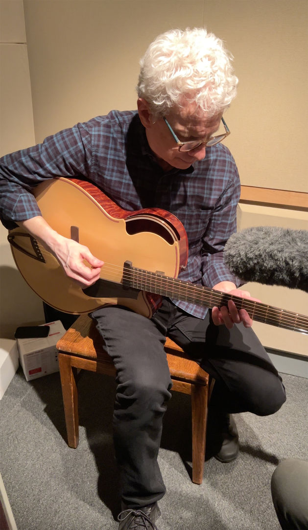 A white-haired man in a blue plaid shirt is seated and playing a guitar. Part of a boom is just visible on the right side of the frame.