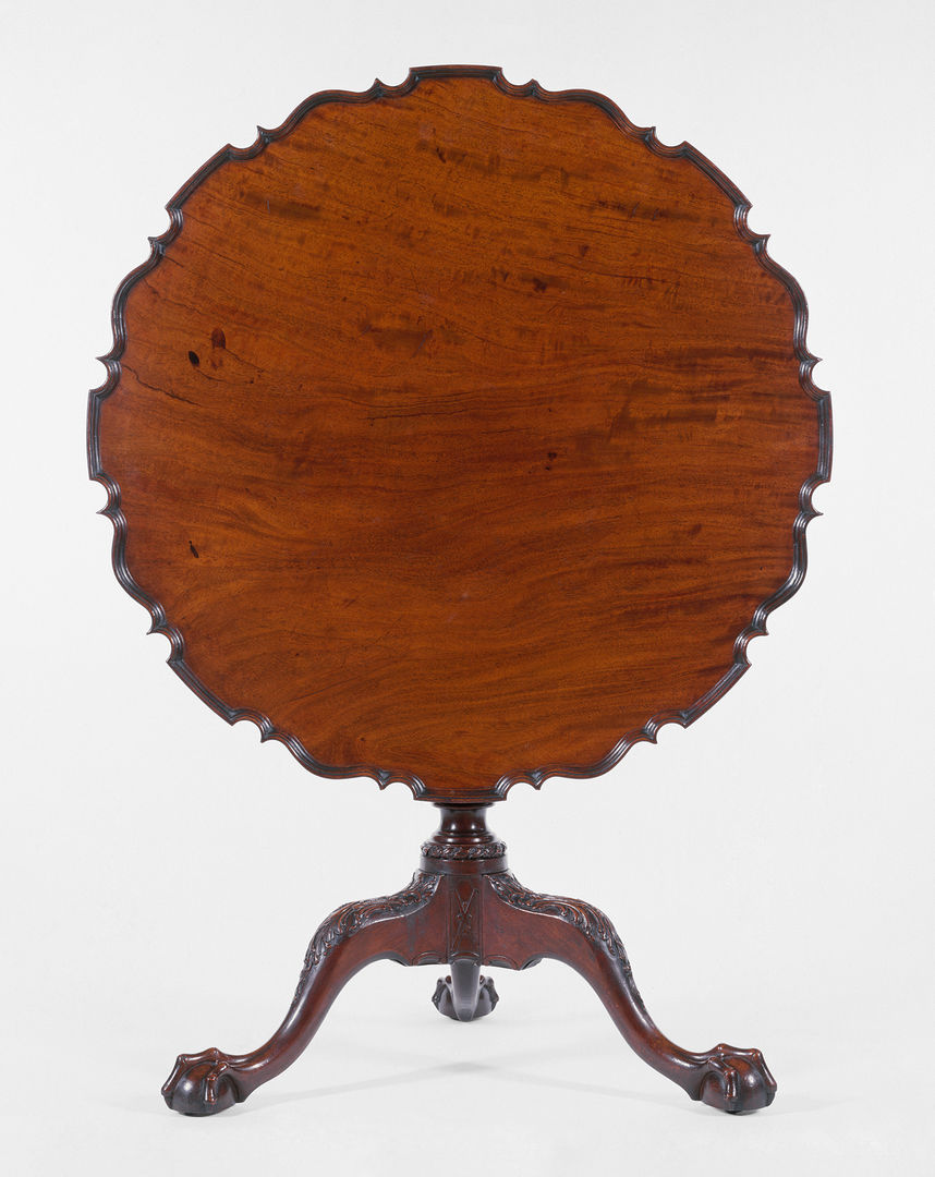 Eighteenth-century mahogany table. The top is tilted upwards ninety degrees, showcasing the scalloped and straight edges of the tabletop. The tripod base is elegantly carved.