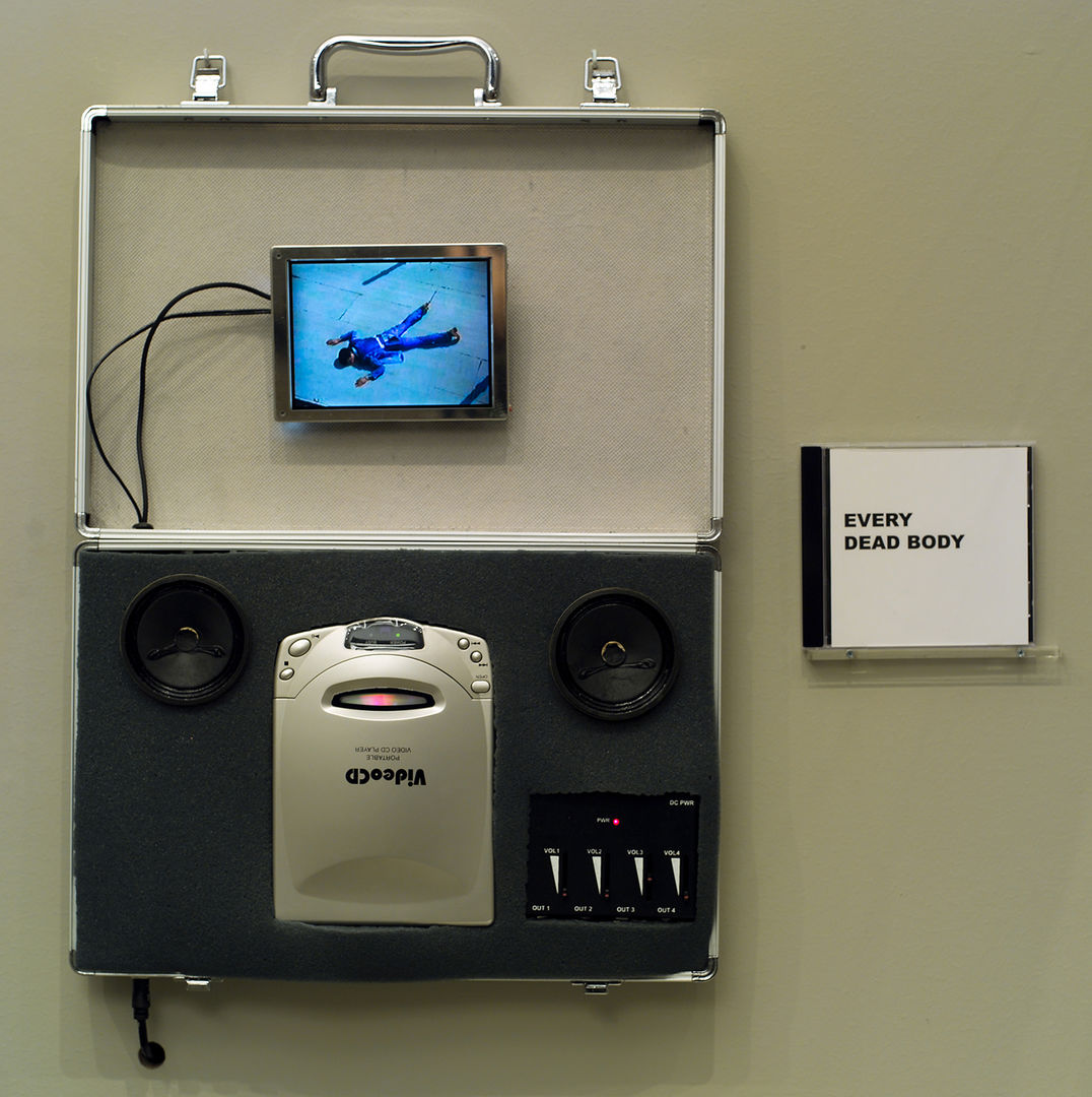 A video CD player with exposed wires, attaching to a small screen showing a figure in blue lying face down. To the right of the player is a small CD with cover art that reads "EVERY DEAD BODY."