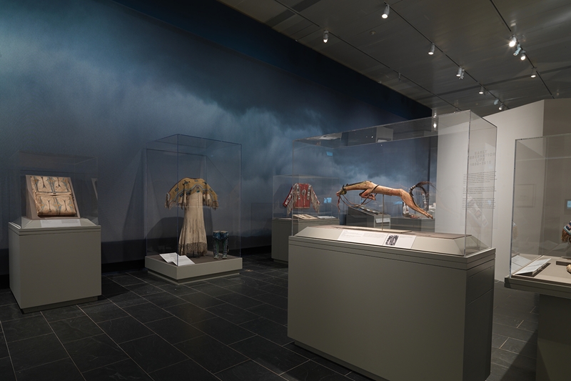 Gallery view from Plains Indians. Indigenous artworks are displayed in glass cases against a wallpaper of blue clouds.