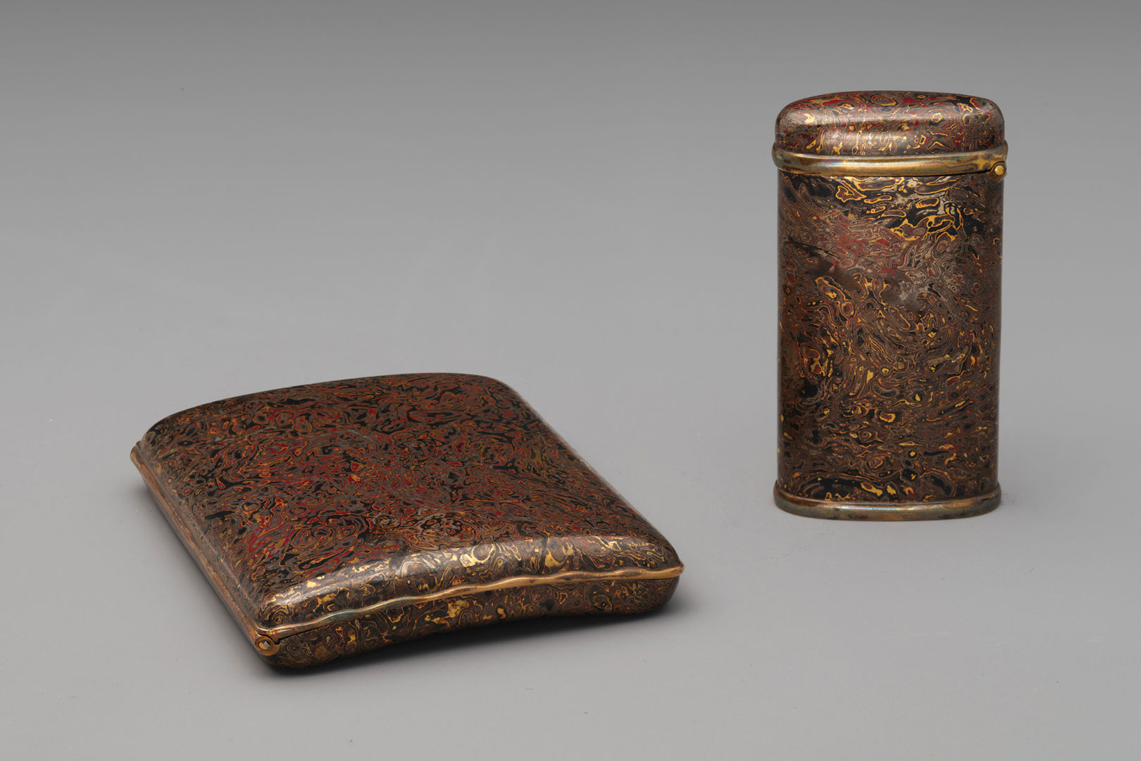 A flat cigarette case and an upright tall match case in a matching wood grain pattern rendered in deep red, black, brown, and gold metals.
